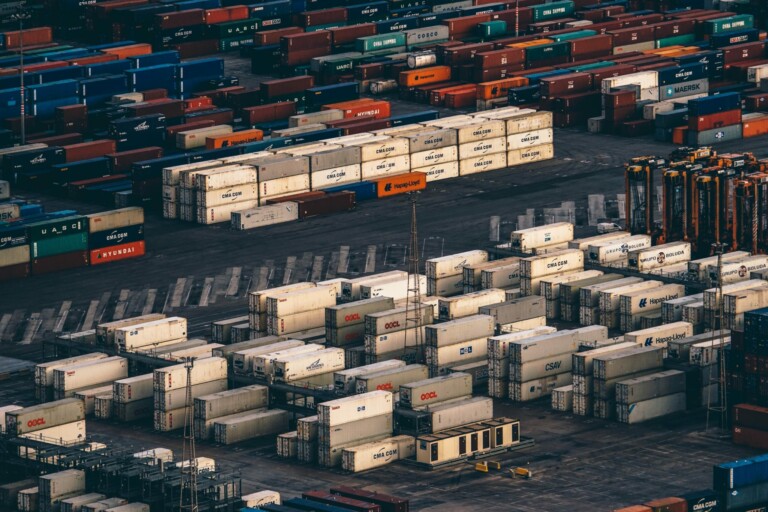 Aerial view of a large shipping yard with numerous cargo containers organized into rows. The containers, many marked with company logos, come in various colors. The industrial setting features stacked containers and structures, emphasizing global trade and the import/export of goods from Australia.