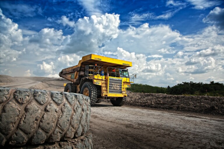 Un gran camión volquete amarillo de minería circula por un camino de tierra en un sitio minero, que recuerda a los del sector minero latinoamericano 2019. En primer plano dominan neumáticos gigantes y arriba se extiende un cielo azul parcialmente nublado. El paisaje árido muestra a lo lejos toques de verdor.