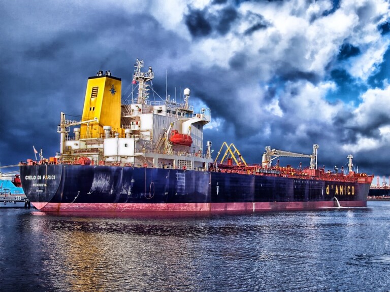 A large cargo ship named "Cielo di Parigi" is docked in a harbor, its red and black hull contrasting against the yellow superstructure. Various equipment adorns its deck. The cloudy, dark sky hints at an impending storm, setting an ominous scene in the Kingdom of The Netherlands' bustling port.