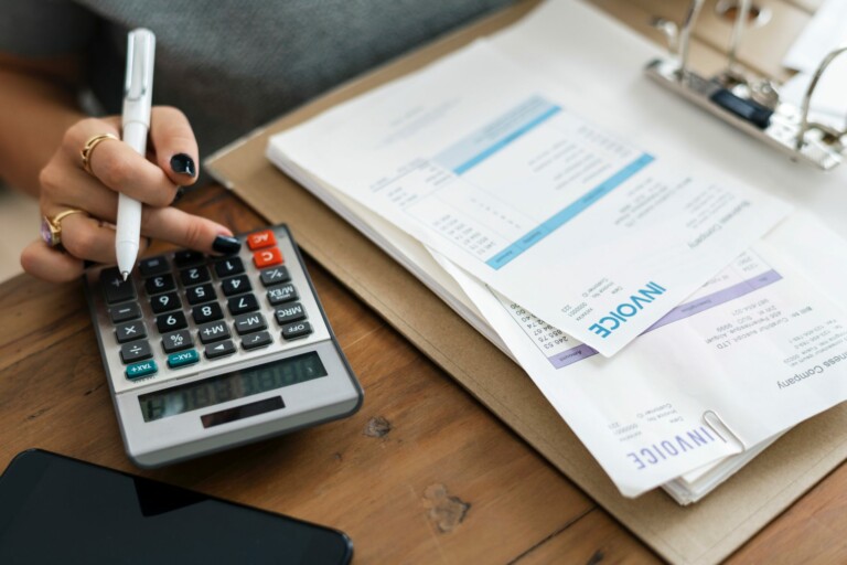 Primer plano de una persona que utiliza una calculadora mientras sostiene un bolígrafo, con varias facturas en un portapapeles y algunas esparcidas sobre la mesa. Un teléfono inteligente es parcialmente visible en la esquina inferior izquierda, lo que refleja la naturaleza meticulosa de Contabilidad e Impuestos en Uruguay. El entorno parece ser un espacio de trabajo para tareas financieras.