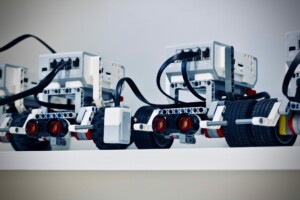 A row of robotic vehicles, constructed with white plastic blocks and various electronic components, lined up on a white surface. Booming with innovation, these Costa Rica-inspired robots feature black wheels, cables, and visible red sensors on the front.