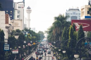 A busy urban street scene features heavy vehicle and motorcycle traffic. Lined with tall buildings, street lamps, and palm trees, the street is packed with movement. A distinctive white tower with a large dome is visible in the background, hinting at burgeoning Indonesian trade opportunities.