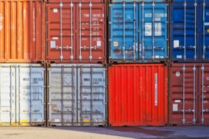 The image shows a stack of shipping containers in various colors, including red, blue, and gray. They are arranged in a three-by-three grid. Labels and numbers are visible on the doors, indicating identification and specifications crucial for tracking import export taxes in Colombia.