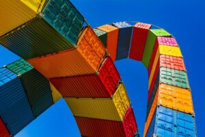 An installation of brightly colored shipping containers, reminiscent of the vibrant economic boost investments brought by the Peru-China FTA, stacked and curved into an archway against a clear blue sky. The containers in red, orange, yellow, green, and blue reflect the dynamic spirit of free trade agreements.