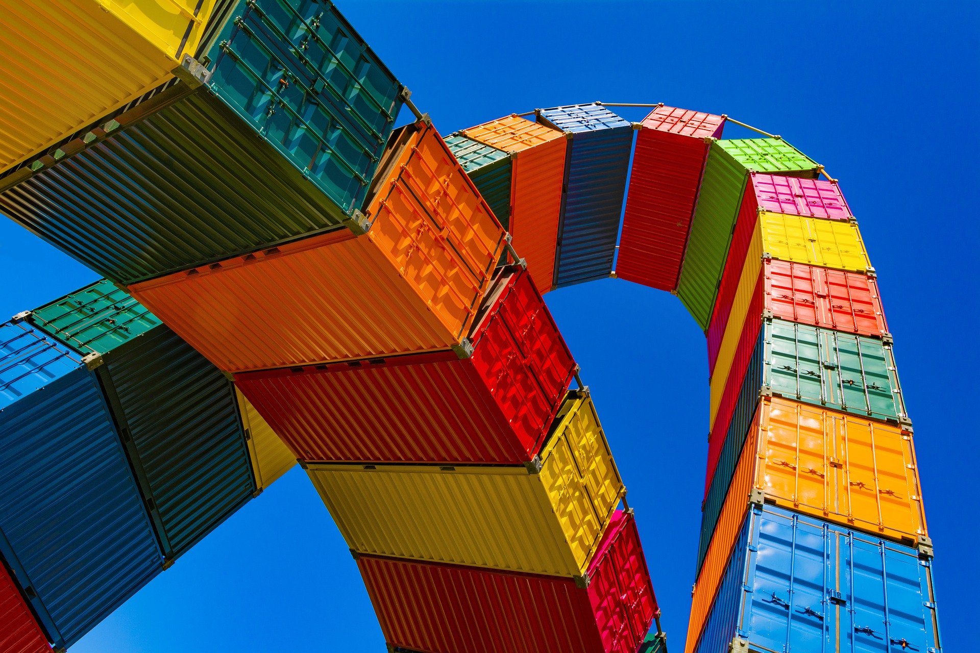 Uma instalação de contêineres coloridos, que lembram os investimentos de impulso econômico vibrante trazidos pelo TLC Peru-China, empilhados e curvados em um arco contra um céu azul claro. Os contêineres em vermelho, laranja, amarelo, verde e azul refletem o espírito dinâmico dos acordos de livre comércio.