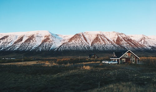 A small house with a tall peaked roof and two parked vehicles sits in a vast, open area with scattered vegetation. In the background, towering, snow-capped mountains rise under Chile's clear blue sky.