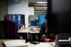 Two people sitting at desks working on computers with screens displaying code in a booming office environment. One person has pink hair, and the other person has headphones on. There are multiple monitors and laptops on the desks, showcasing how vibrant Costa Rica's service sector can be.