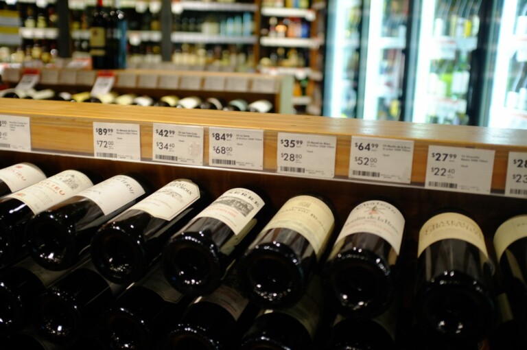 A liquor store display with multiple shelves of wine bottles. The front row shows various wine bottles laid horizontally. Above the bottles, price tags and descriptions are visible, displaying prices ranging from $28.80 to $72.29—perfect for those looking to import wine into Mexico.