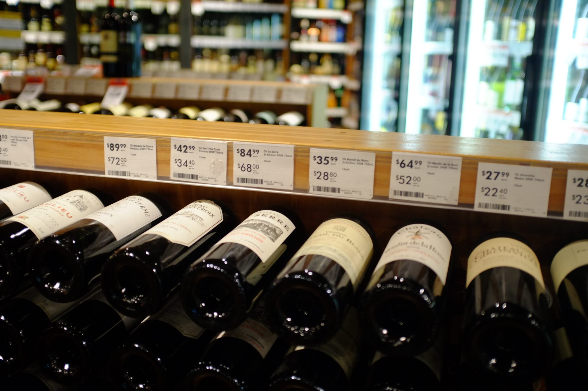 A liquor store display with multiple shelves of wine bottles. The front row shows various wine bottles laid horizontally. Above the bottles, price tags and descriptions are visible, displaying prices ranging from $28.80 to $72.29—perfect for those looking to import wine into Mexico.