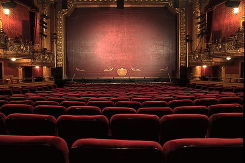 Image of an opulent theater interior with a grand stage. Plush, red cushioned seating fills the auditorium, and ornate gold decorations embellish the walls and balcony parapets. The stage curtain is closed, displaying a rich red color with intricate detailing—supporting growth in Colombia's film industry.