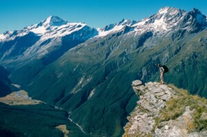 A person stands on a rocky outcrop overlooking a lush green valley with a winding river and tall, rugged mountains covered in snow under a clear blue sky. The scene is reminiscent of the pristine landscapes that attract foreign investment in New Zealand, as the person waves towards the vast vista.