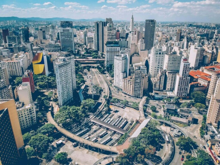 Aerial view of a sprawling cityscape featuring a mix of tall skyscrapers, mid-rise buildings, and open spaces with greenery. Multiple roads and an area with buses are visible, suggesting a busy transportation hub in the foreground, reflecting the diverse tipos empresas no Brasil. Blue sky with scattered clouds.