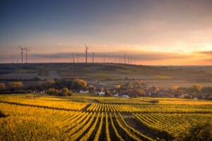 A landscape at sunset in Colombia features a series of wind turbines on a distant hill. In the foreground, there are neatly arranged rows of crops in a field. A small cluster of houses is situated between the fields and the turbines, highlighting unique renewable energy business opportunities. The sky is partly cloudy.