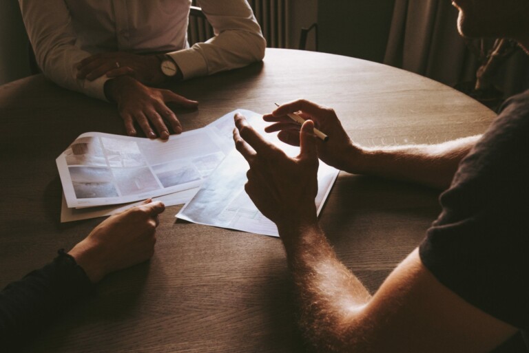 Three people are seated around a wooden table engaged in a discussion. They have documents spread out on the table. Two individuals' hands are visibly holding and pointing at the papers, while the third person's hand rests on the table. The papers appear to contain details about a Free Trade Agreement between Australia and Peru.