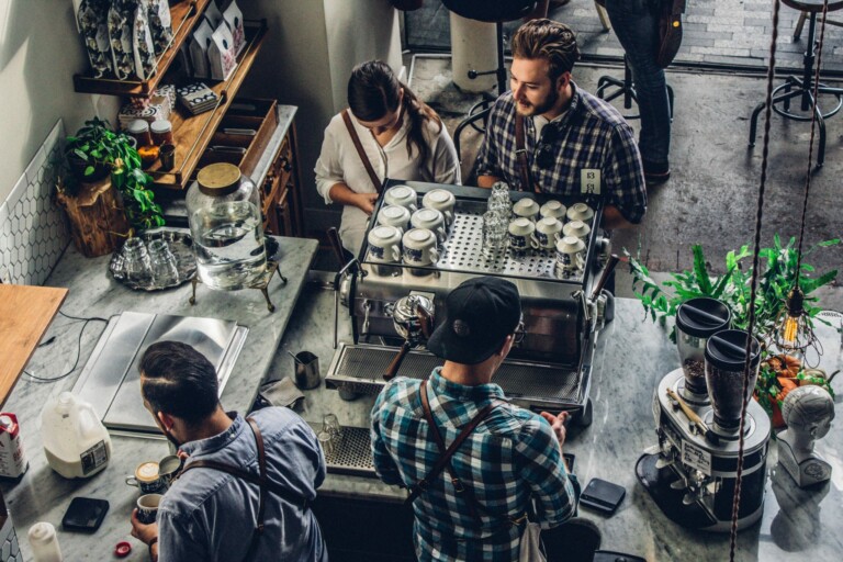 A top-down view of a coffee shop counter with four people. Two baristas, one wearing a black cap and the other with long hair, are preparing drinks. Two customers wait, one looking at the counter and the other using a smartphone. Various coffee equipment from Empresa is visible around them.