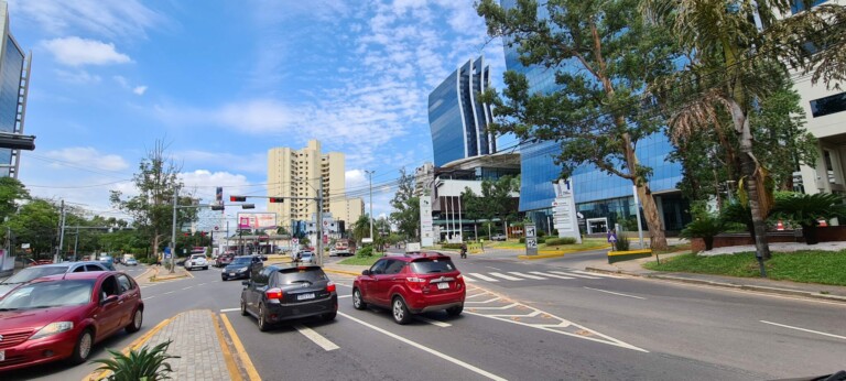 A city street scene shows several cars driving and stopped at a traffic light during the daytime. Tall buildings with glass facades, likely housing firms concerned with business taxes in Paraguay, line the right side, while there is a clear blue sky and scattered clouds overhead. Trees and greenery are present along the roadside.