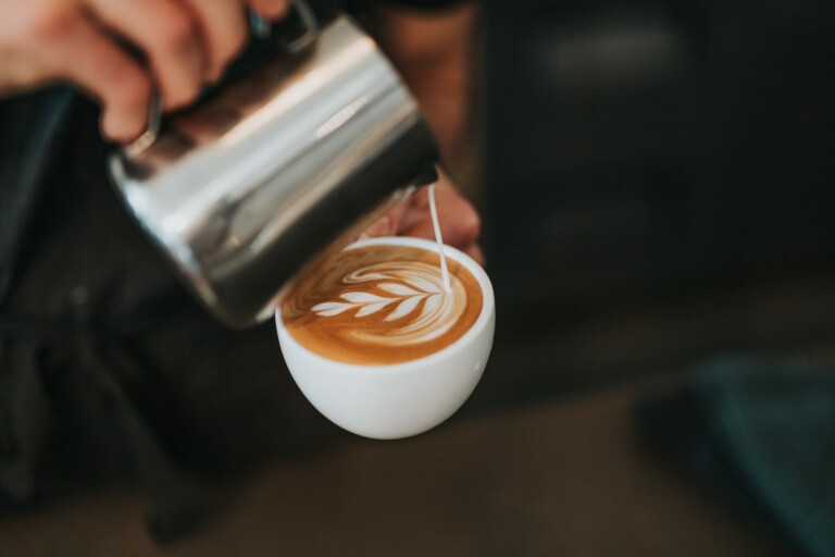 A close-up of a hand pouring steamed milk from a metal pitcher into a cappuccino cup, creating latte art on the surface of the coffee. The design is a leaf pattern, showcasing the rich coffee culture in Australia influenced by LatAm exporters.