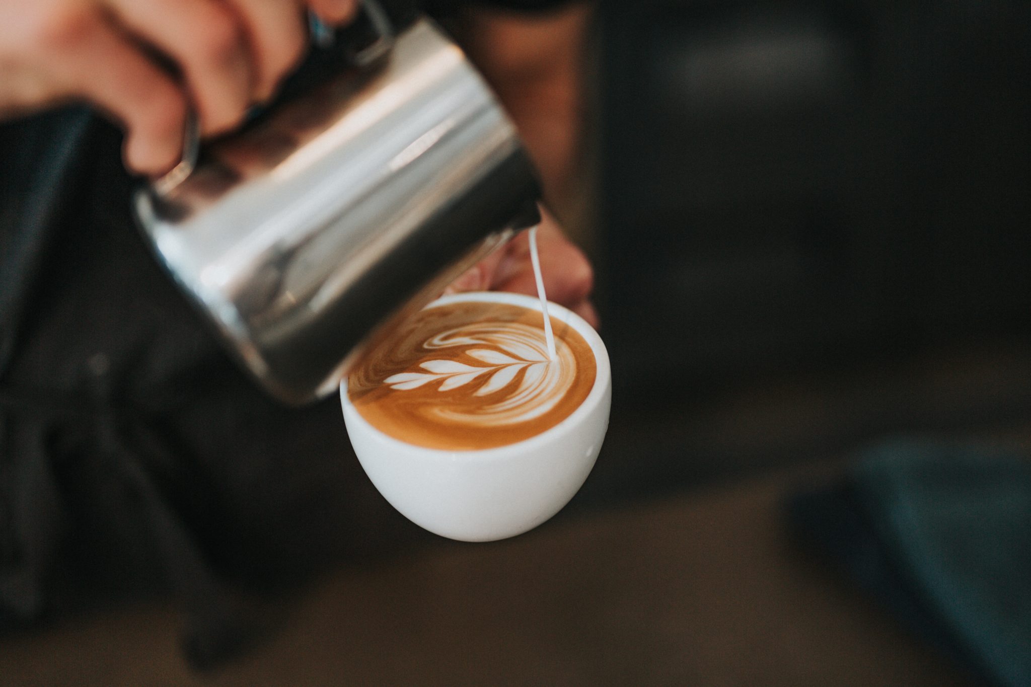 Um close-up de uma mão despejando leite vaporizado de uma jarra de metal em uma xícara de cappuccino, criando latte art na superfície do café. O design é um padrão de folhas, mostrando a rica cultura do café na Austrália, influenciada pelos exportadores da América Latina.