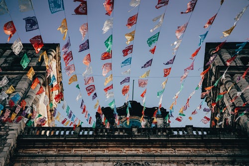 Colorful flags are strung across the sky between two stone buildings showcasing detailed architecture, including columns and arches. Amidst discussions of Australian Foreign Direct Investment, the festive flags stand out against a clear blue sky from a perspective below, looking upward.