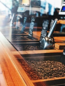 A coffee shop counter features several trays filled with coffee beans and metal scoops, showcasing the vibrant Coffee Culture. Small blackboards with white text provide information about different coffee varieties from LatAm Exporters. The background shows a blurred scene of the bustling coffee shop interior.