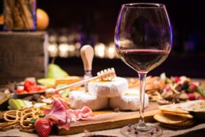 A glass of red wine is placed on a table alongside a wooden board filled with various snacks. The board includes cheese, crackers, prosciutto, strawberries, and a honey dipper. The background is softly lit, highlighting the food and drink—a perfect setting to appreciate LatAm Exporters' finest offerings.