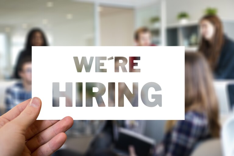 A person is holding a white sign with the cutout words "WE'RE HIRING EMPLOYEES IN AUSTRALIA." In the background, a group of people is sitting and standing in a modern office setting, partially visible through the cutout letters of the sign.