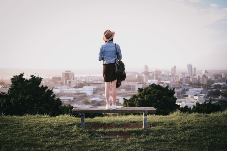 A person stands on a bench on a grassy hilltop, gazing at a cityscape in the distance. They are wearing a hat, a denim jacket, a black skirt, and white sneakers, and are carrying a backpack with travel brochures about Visa Types in New Zealand. The background features various buildings and a pale sky.