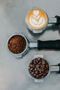 Three portafilters arranged vertically against a gray background. The top one holds a cup of latte art coffee, showcasing Coffee Culture at its finest; the middle one contains ground coffee, and the bottom one is filled with whole beans ready for LatAm Exporters.