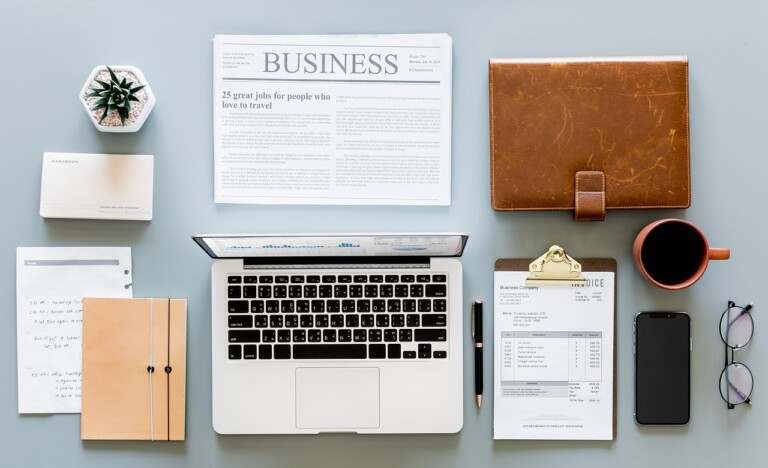 Un espacio de trabajo con una computadora portátil, una libreta de cuero, una pila de papeles, una taza de café, un par de vasos, un teléfono inteligente, una planta, un periódico titulado "Business", tarjetas de presentación, un bolígrafo y un portapapeles con documentos comerciales .