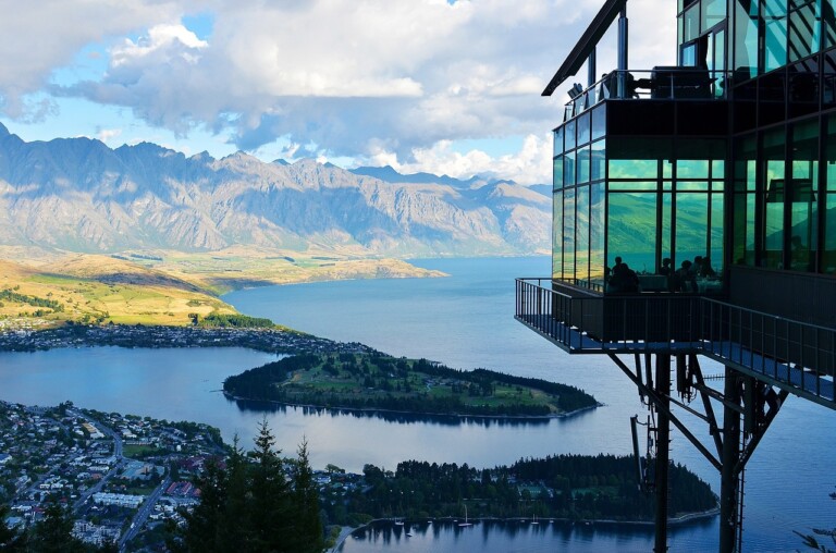 A modern building with large glass windows extends from a hillside overlooking a scenic landscape. Below, there is a lake with an island, a town, and distant mountains under a partly cloudy sky. Inside the building, where people are enjoying the view, business discussions about Formação da Empresa take place.