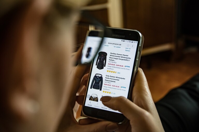 A person is holding a Samsung smartphone and shopping for jackets on Amazon. The screen displays images and details of different jackets with ratings and prices, showcasing the benefits of e-commerce. The person's fingers touch the screen, while another hand is partially visible holding the phone.