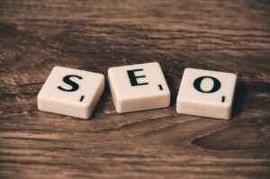 Three letter tiles spelling out "SEO" are placed on a wooden surface. The tiles are white with black letters and each have a small numerical value in the corner, resembling Scrabble tiles—a nod to the importance of technology in effective digital strategies.