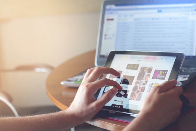 A person uses a tablet to browse a website displaying various images, while a laptop in the background shows an email inbox. Both devices, symbolizing the rise of technology in Peru, rest on a wooden table in a softly lit room.