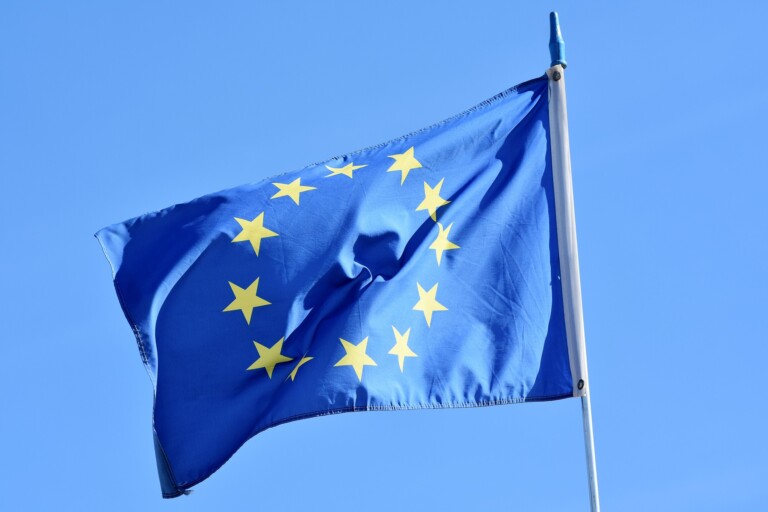 A blue flag featuring twelve yellow stars arranged in a circle, symbolizing EU cooperation, is waving on a flagpole against a clear blue sky.