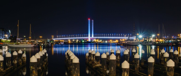 A nighttime view of a harbor with docked boats and illuminated poles in the foreground. In the background, a bridge with vertical columns is lit up, reflecting on the calm water. City lights are visible, enhancing the scene's brightness against the dark sky—truly one of the key outcomes of urban beauty.