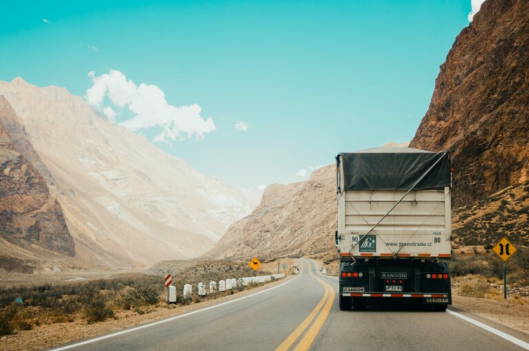 A white cargo truck, possibly serving mining operations, drives down a winding road through a mountainous desert landscape under a blue sky. Road signs and barriers are visible on the roadside, and tall, rocky cliffs rise on both sides of the road in Ecuador.