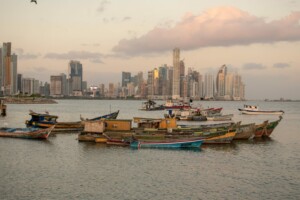 Fishing boats are anchored in the foreground, with a city skyline featuring tall buildings in the background. The sky is partly cloudy with a soft, pastel-colored sunset, and a bird is visible flying in the upper left corner of the image, capturing the essence of Central America's coastal beauty.