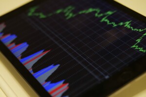 A close-up image of a tablet screen displaying financial graphs related to mining in Bolivia. The screen shows a grid with green line graphs at the top and multi-colored bar charts in blue, red, and gray at the bottom, indicating various financial data trends.