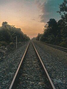 A railway track stretches into the distance, flanked by dense vegetation on both sides. The sky is a mix of warm hues, indicating either sunrise or sunset, with scattered clouds. The rails and gravel create a perspective that converges towards the horizon, symbolizing the international expansion through Central America.