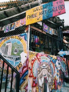 A colorful outdoor art gallery showcases vibrant murals on a staircase. A sign reading "Welcome to Free Art Gallery" hangs above. Various paintings are displayed, with one person doing business in Colombia visible near the entrance. The artwork includes diverse and abstract designs.