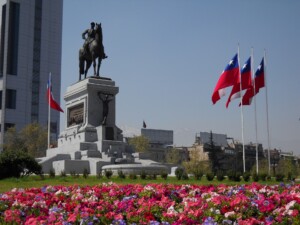 A statue of a person riding a horse stands prominently on a pedestal in a bustling city square in Argentina. Colorful flowers bloom in the foreground, and several flags with a blue, white, and red design are displayed on flagpoles nearby. Tall buildings rise in the background, symbolizing progress driven by Free Trade Agreements.