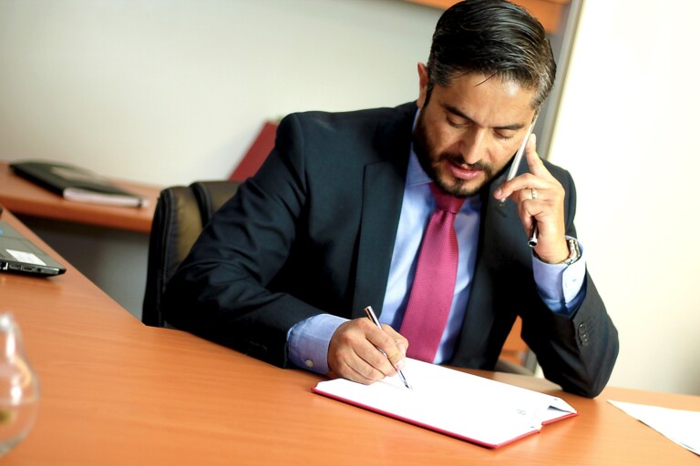 A legal representative in a dark suit and red tie is sitting at a desk, holding a phone to his ear with one hand while writing on a document with the other. The desk has a few papers and a closed laptop. He appears to be in an office setting.