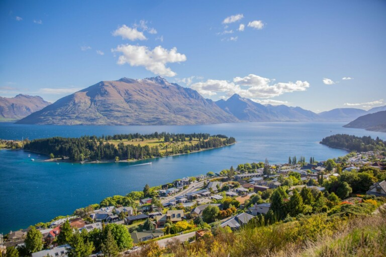 A scenic view of a lake surrounded by mountains and a small town illustrates the peaceful synergy often found in business in New Zealand. The blue, calm lake with green islets mirrors the town's buildings and houses nestled among trees, under a clear sky dotted with scattered clouds.