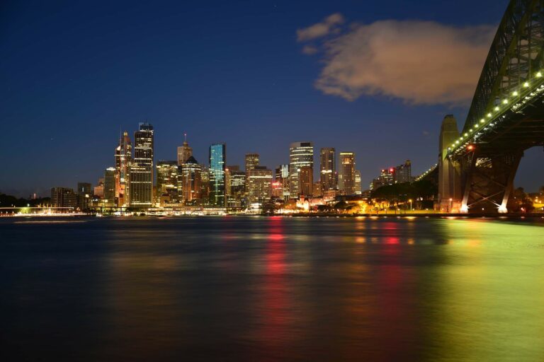 A nighttime cityscape shows a skyline with numerous illuminated buildings beneath a clear, dark blue sky. A brightly lit bridge extends from the right side of the image, and multicolored reflections from the lights are visible on the water below, much like how a legal representative in Australia navigates through complex cases.