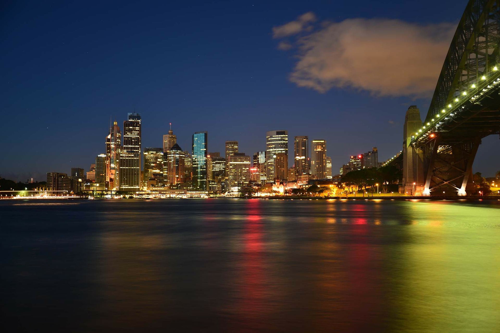 Uma paisagem urbana noturna mostra uma linha do horizonte com vários edifícios iluminados sob um céu azul escuro e claro. Uma ponte bem iluminada se estende do lado direito da imagem, e reflexos multicoloridos das luzes são visíveis na água abaixo, de forma muito parecida com a maneira como um representante legal na Austrália navega em casos complexos.