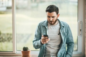 Un hombre con barba está apoyado en el alféizar de una ventana, mirando su teléfono, probablemente navegando por el comercio electrónico en Nueva Zelanda. Lleva una camiseta gris con un logotipo y una camisa vaquera azul claro. Hay un pequeño cactus en maceta en el alféizar de la ventana junto a él.