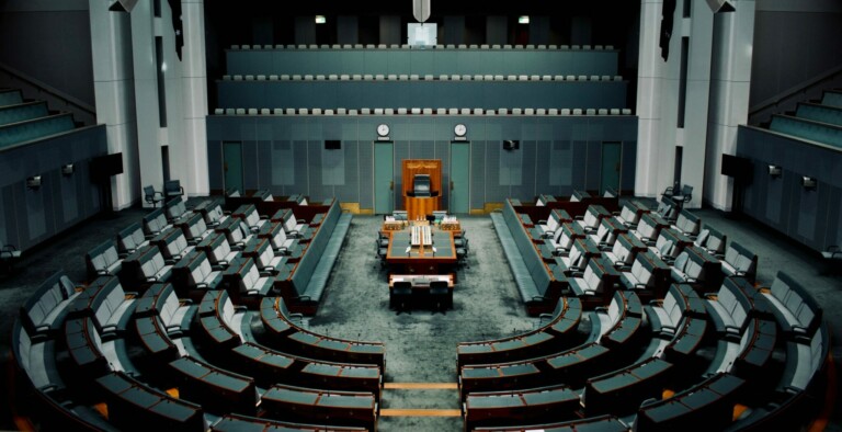 A modern legislative chamber with green seating arranged in a semicircular fashion, facing a central speaker's podium where trade agreements are often discussed. The room is symmetrical with upper balconies and tall white pillars on each side. The floor is carpeted, and the walls are gray with small windows.