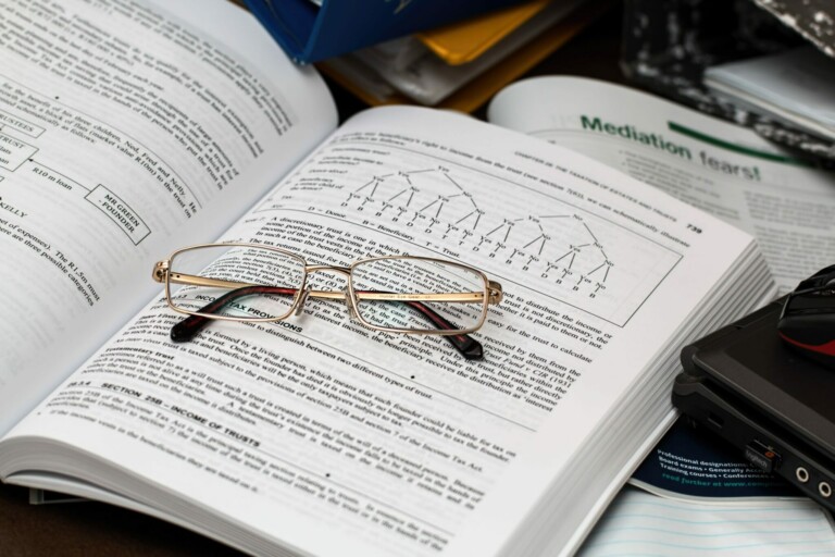 A pair of eyeglasses rests on an open book with text and diagrams related to entity health check mediation. The book is surrounded by other study materials, including a laptop and various papers. The scene suggests a study or work environment.