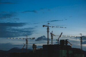 Silhouetted construction cranes stand against a twilight sky. In the background, the outline of partially constructed buildings is visible, while distant mountains can be seen on the horizon. The scene captures the essence of investing in Mexico's booming infrastructure during dusk.