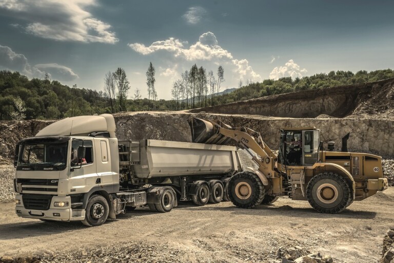 A construction site where a large yellow front loader is transferring materials into the trailer of a white dump truck. The background features rocky terrain and trees under a partly cloudy sky, hinting at the involvement of mining service companies managing the site.